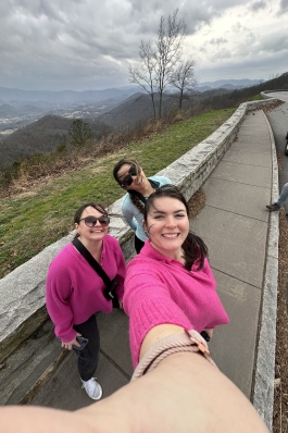 Eve taking a selfie with a group of people, with stunning mountains and a cloudy sky in the background.
